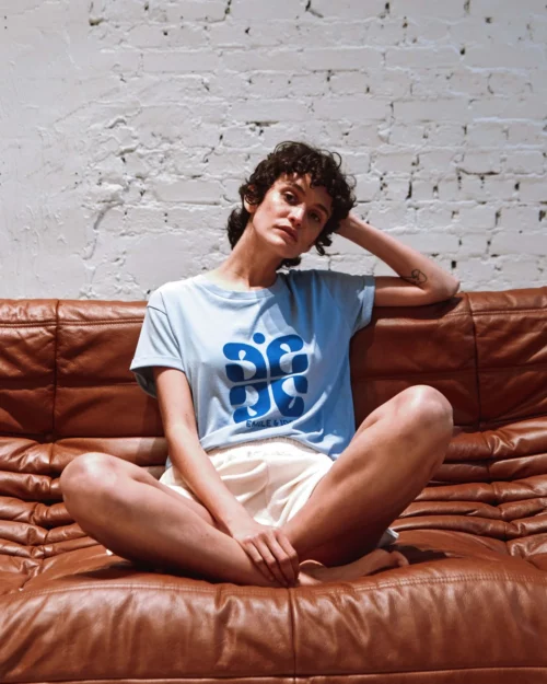 Femme portant un t-shirt bleu ciel en coton bio Emile et Ida avec un imprimé bleu, assise sur un canapé en cuir.