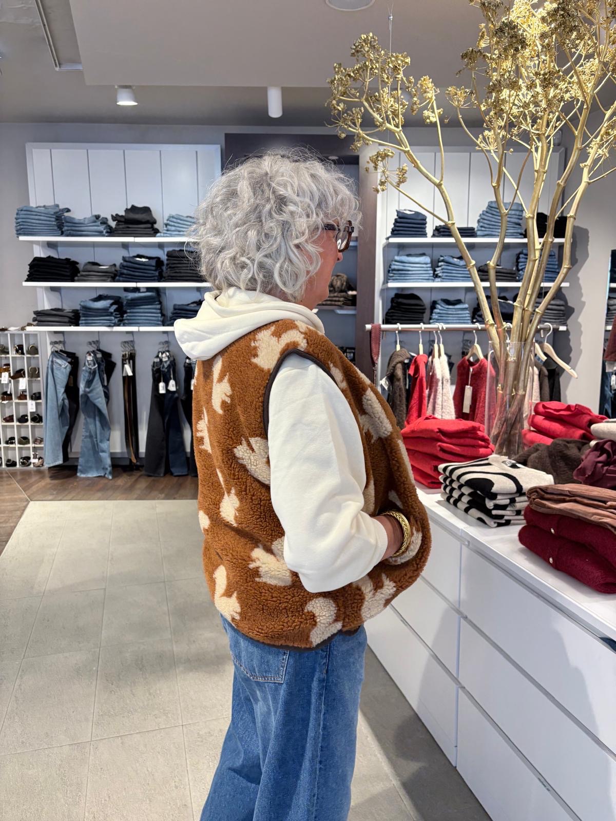 Vue de côté du look femme automne hiver : gilet sans manches sherpa camel motif écureuil et sweat crème à capuche, associé à un jean large bleu, style décontracté et tendance.